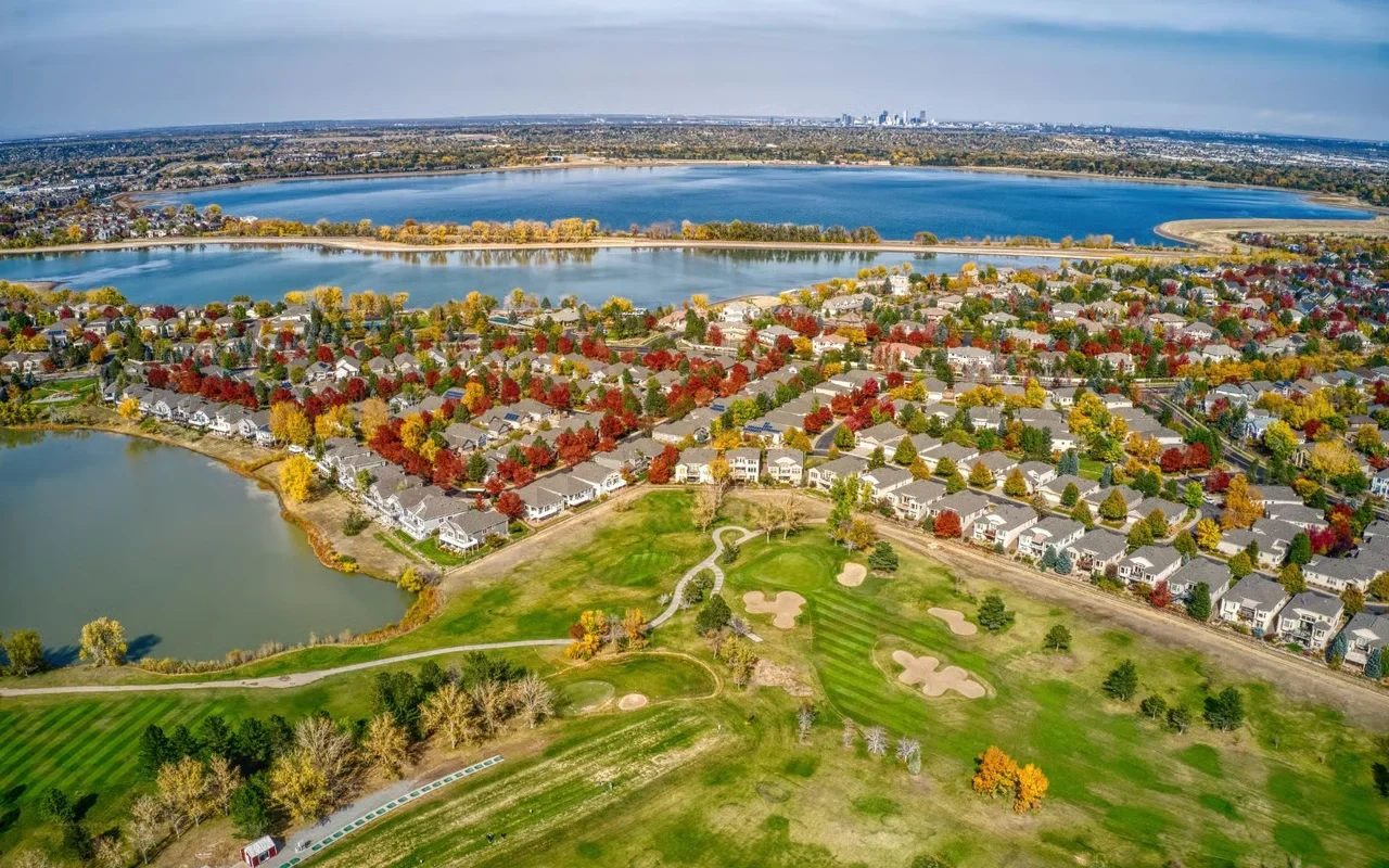 Aerial view of a suburban neighborhood with a golf course in the foreground, a lake, and a city skyline in the background during autumn.
