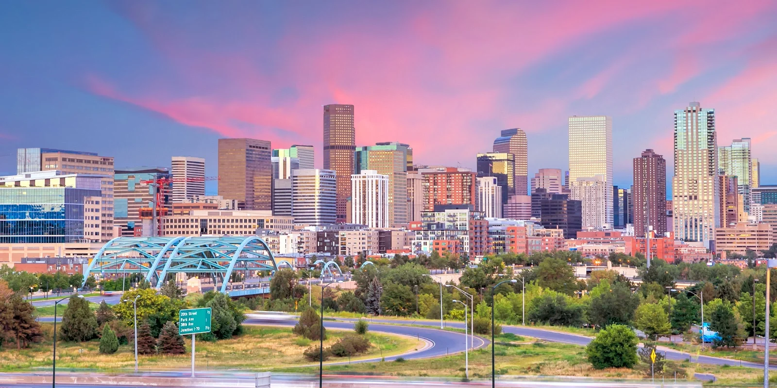 City skyline of Denver, Colorado at sunset with skyscrapers, a park in the foreground, and a pink and purple sky.