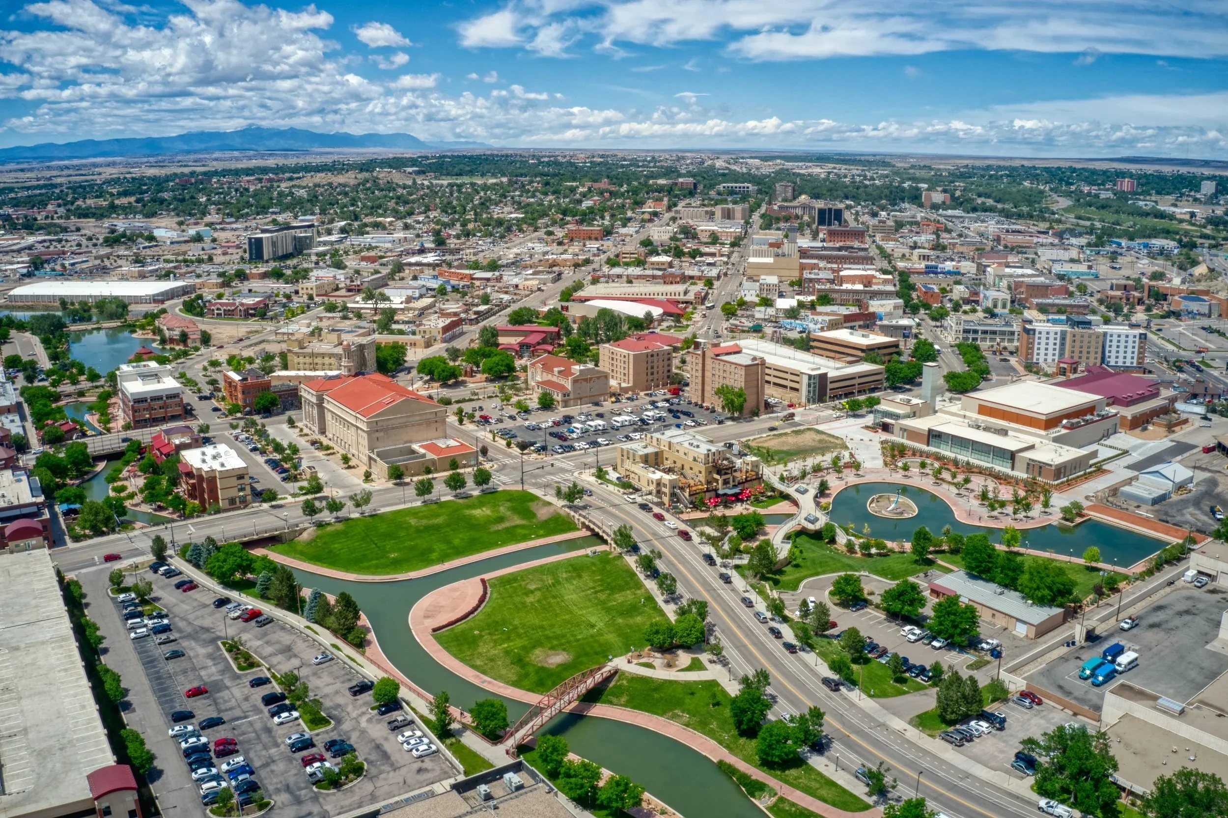 Aerial view of a cityscape with parks, buildings, roads, lakes, and a fountain, under a partly cloudy sky.