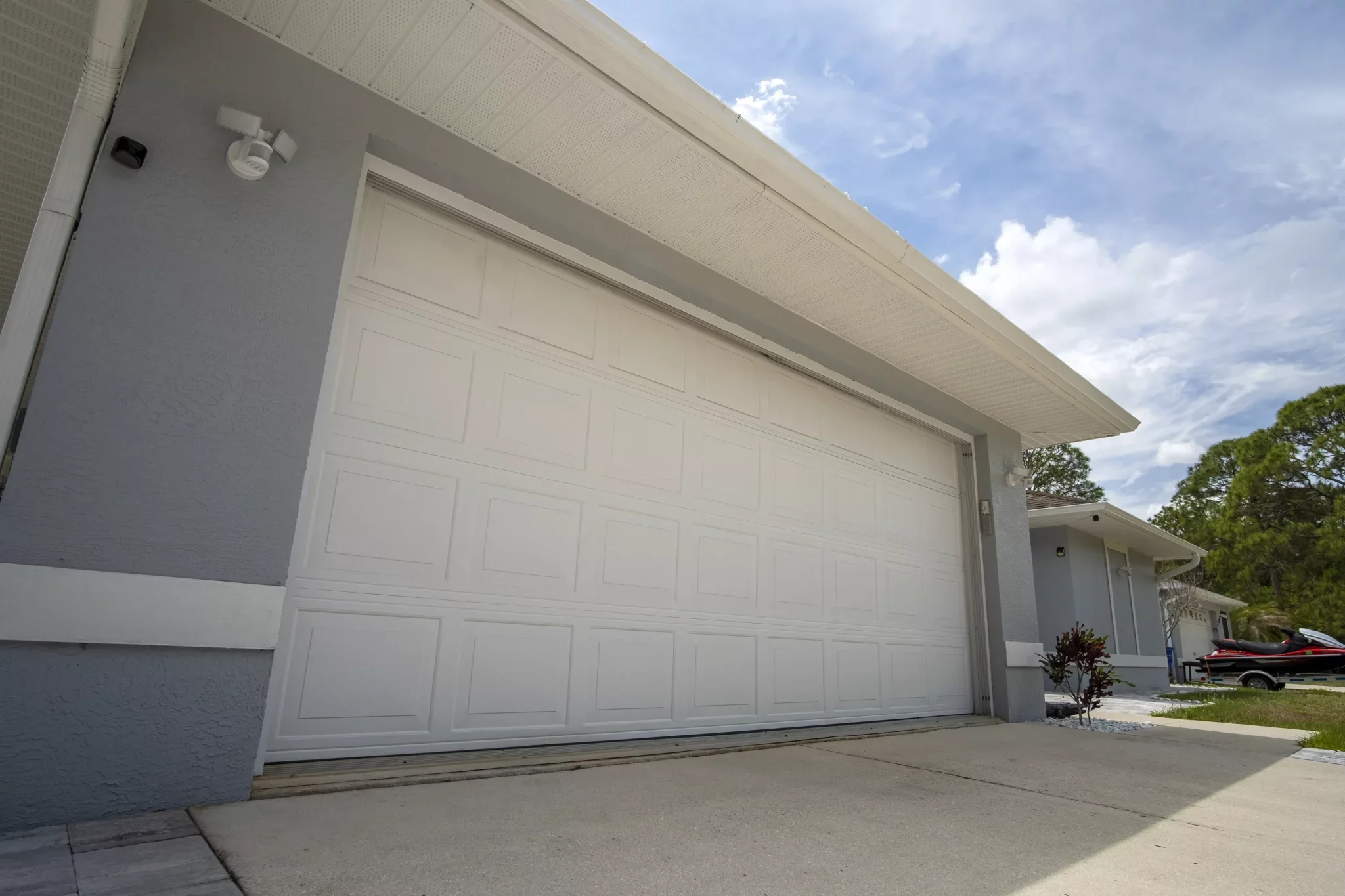 A white garage door on a modern house with a gray exterior wall, a small plant to the right, and a driveway leading up to it, with a partly cloudy sky in the background.