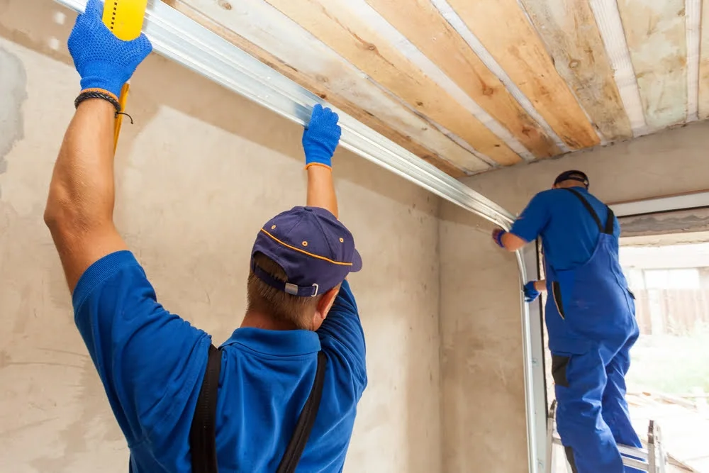 Two workers installing a garage door track in a residential garage, wearing blue uniforms and gloves.