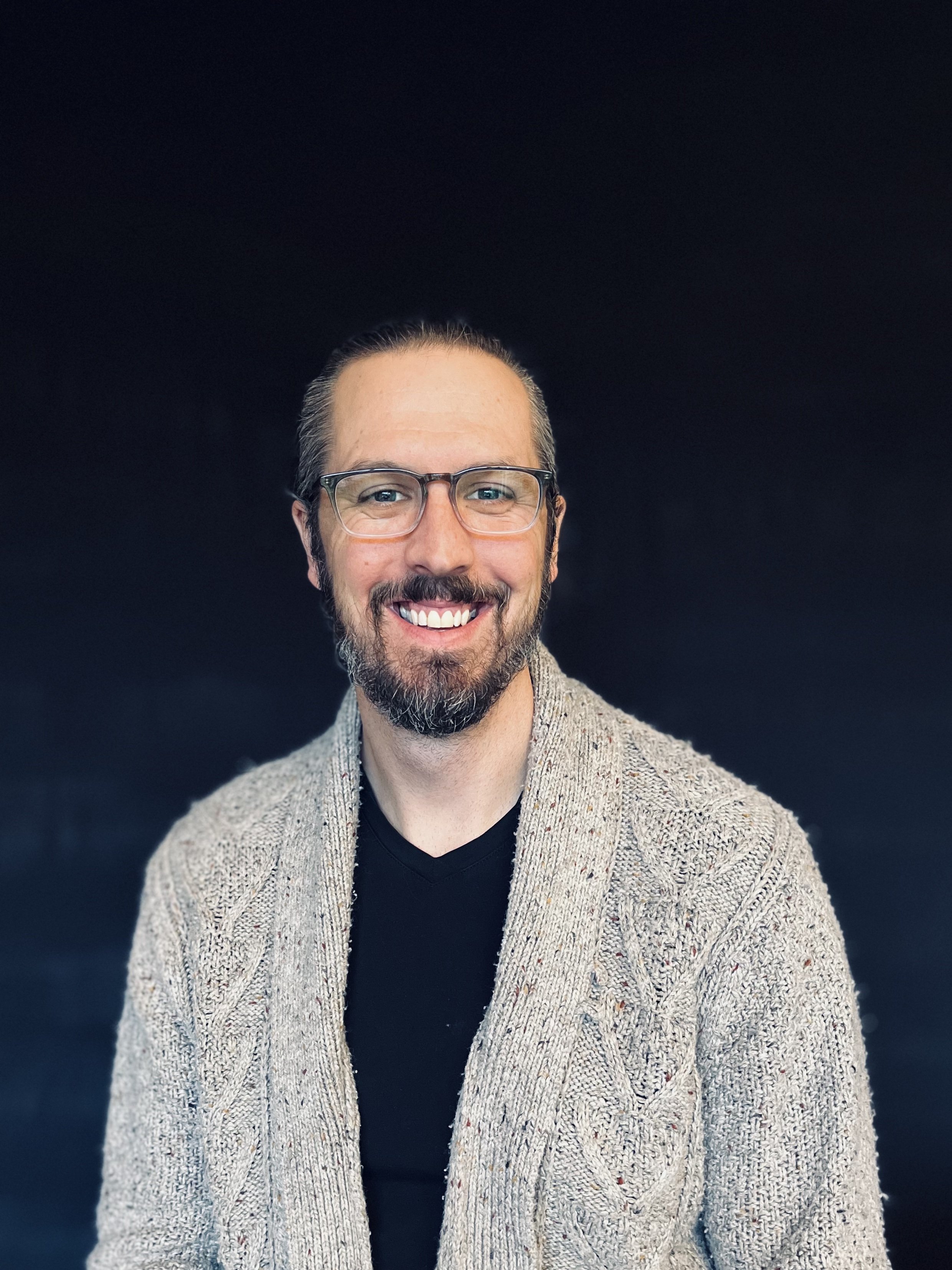 A smiling man with glasses, a beard, and short hair, wearing a patterned cardigan over a black shirt, against a black background.