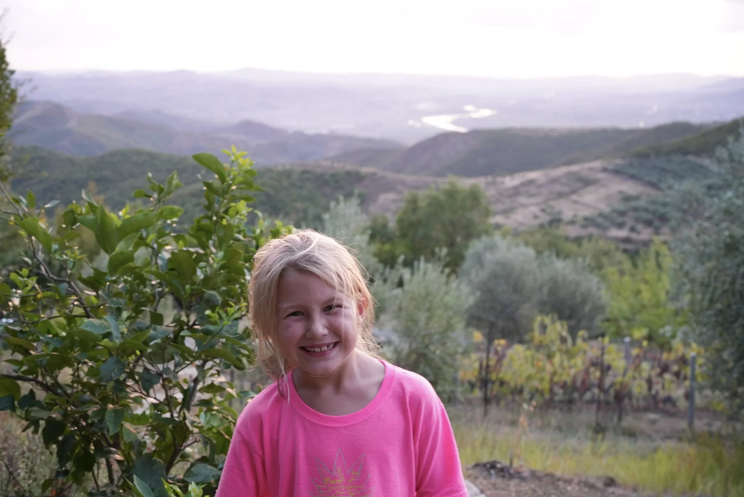 A young girl with blonde hair wearing a pink shirt smiling in a garden with green foliage and distant hills visible in the background.