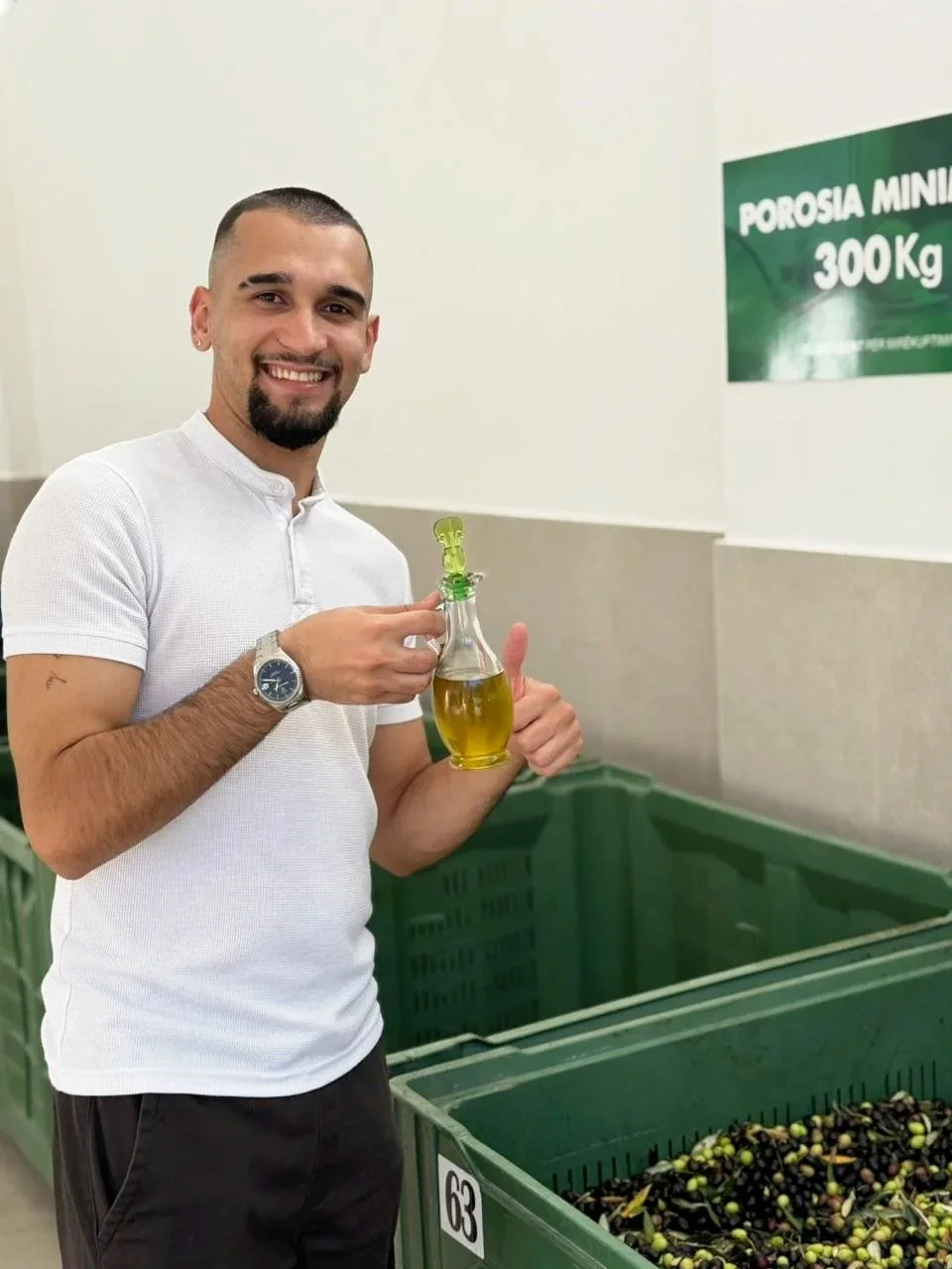 A man with a beard and short hair smiling and holding a glass bottle of yellow oil or liquid, standing next to green crates filled with black olives, in a store or warehouse setting.