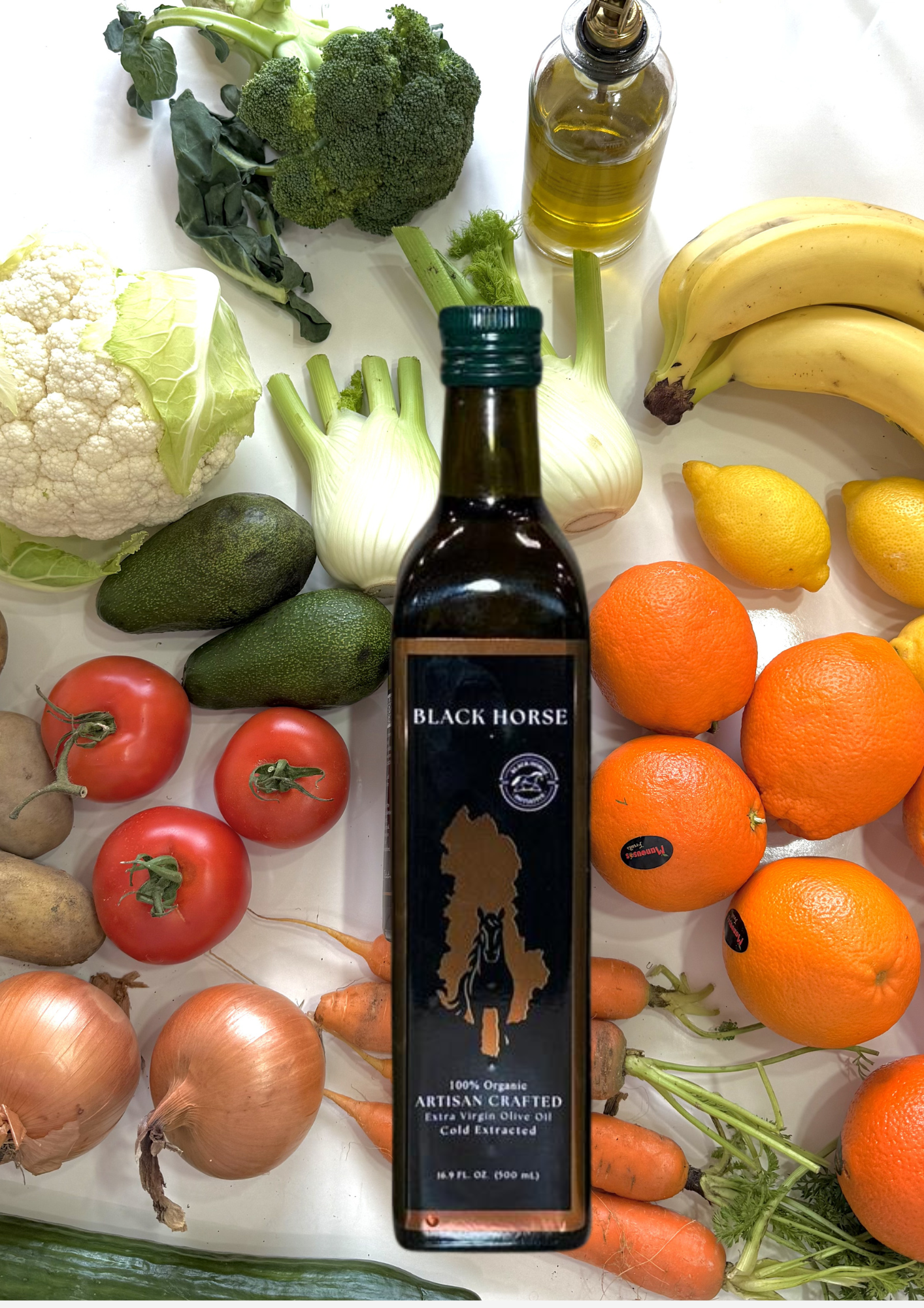 Assorted fresh vegetables, including broccoli, cauliflower, avocados, tomatoes, onions, carrots, and citrus fruits, along with a bottle of Black Horse extra virgin olive oil on a white surface.