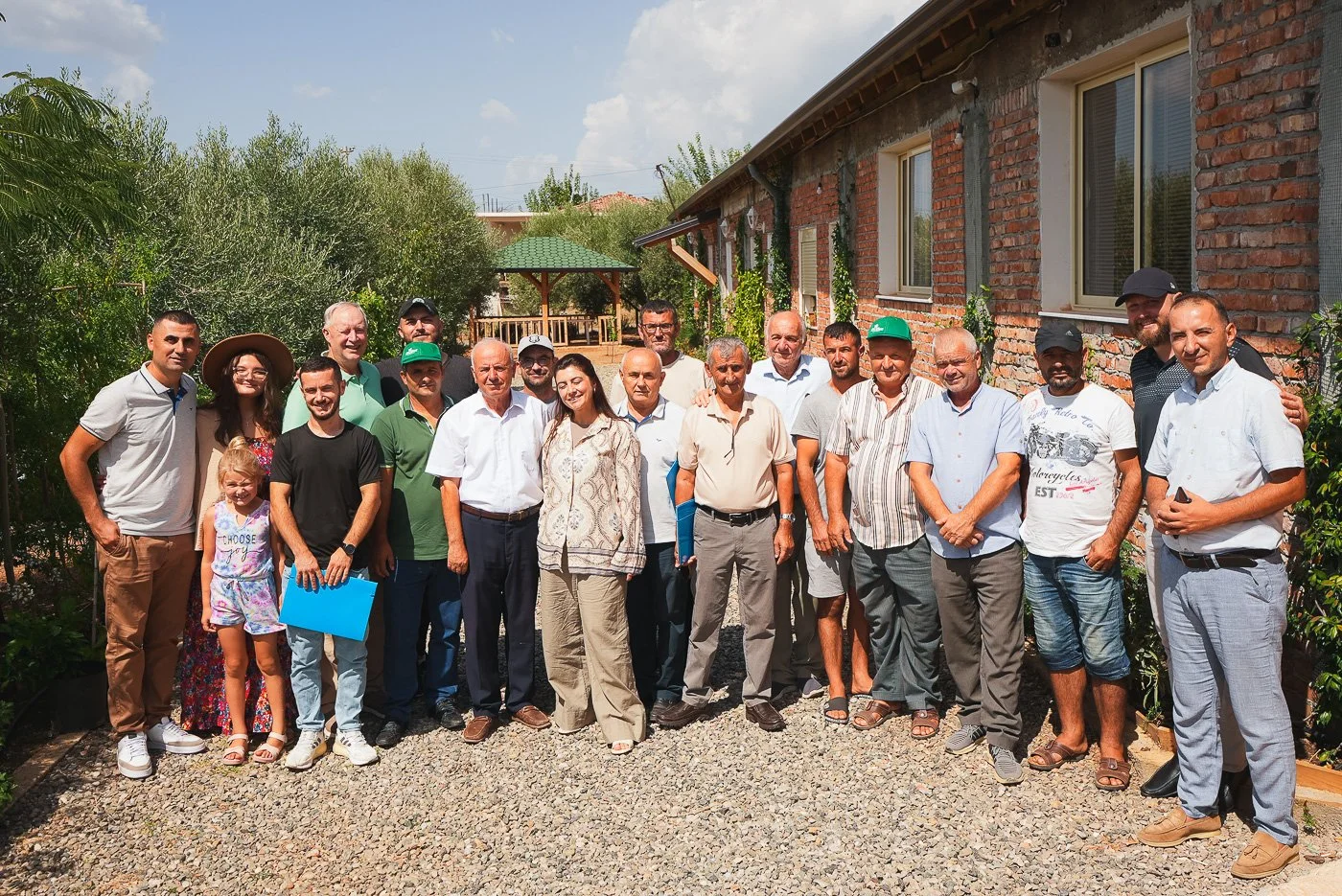 Group of people standing outdoors in front of a brick building with green trees in the background.