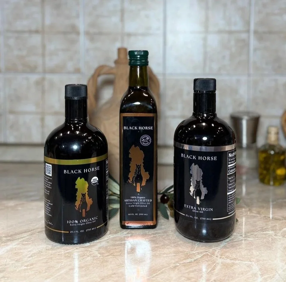Three bottles of Black Horse olive oil on a kitchen countertop with a background of beige tiled wall and a ceramic jug in the background.