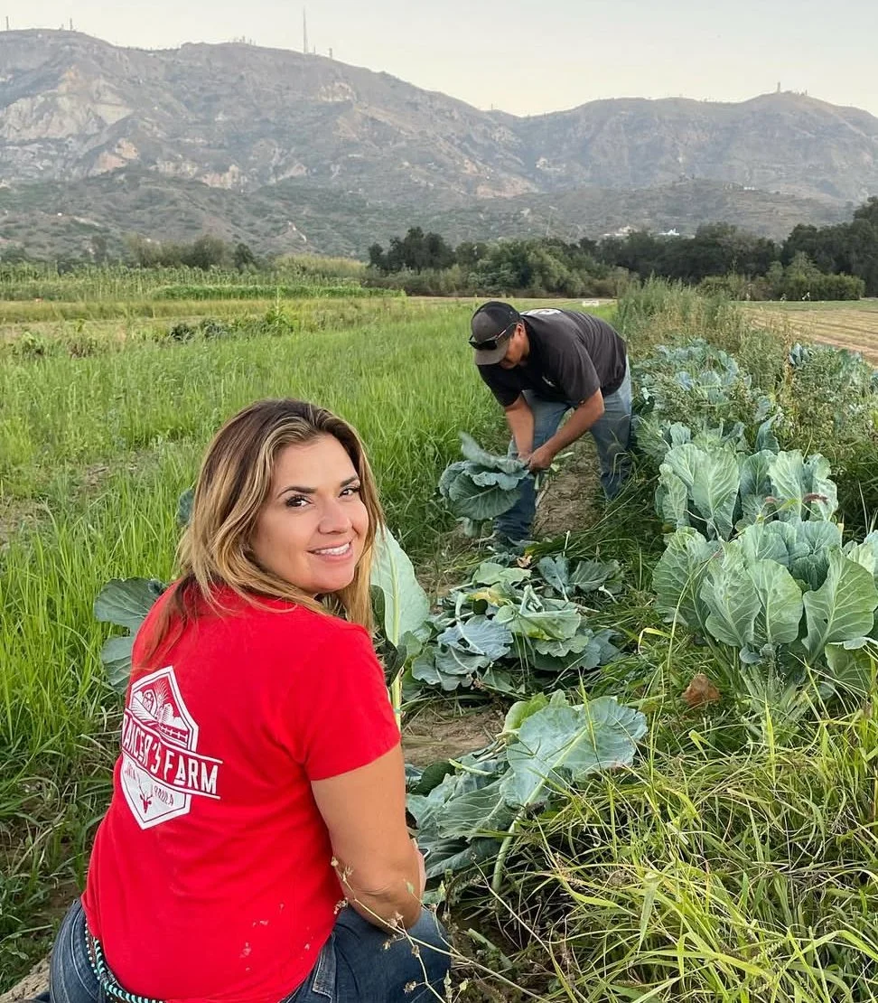 A woman with long blonde hair wearing a red shirt and jeans, smiling at the camera, sitting in a lush green farm field. A man wearing a black shirt, jeans, and a cap is working in the background, tending to the plants, which appear to be cabbages or similar leafy greens. The field is surrounded by rolling hills or mountains in the distance.