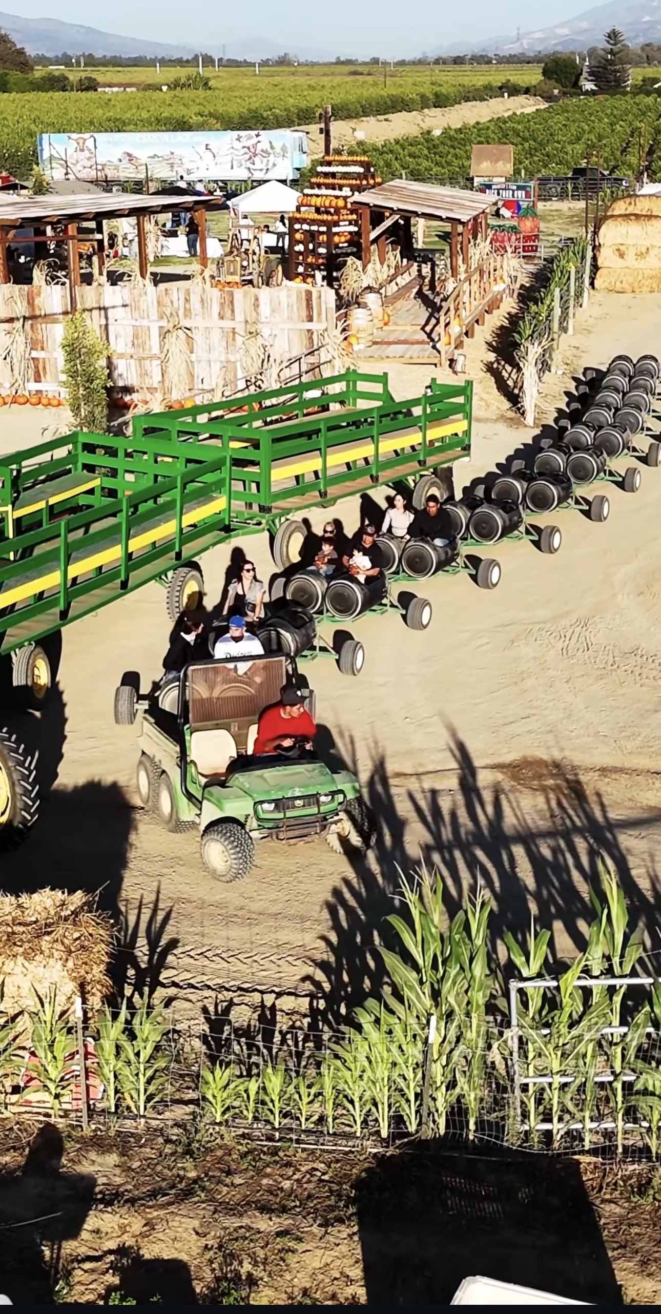 People riding a green golf cart at a farm-themed amusement park, with tractor-shaped cars lined up behind it and farm fields in the background.