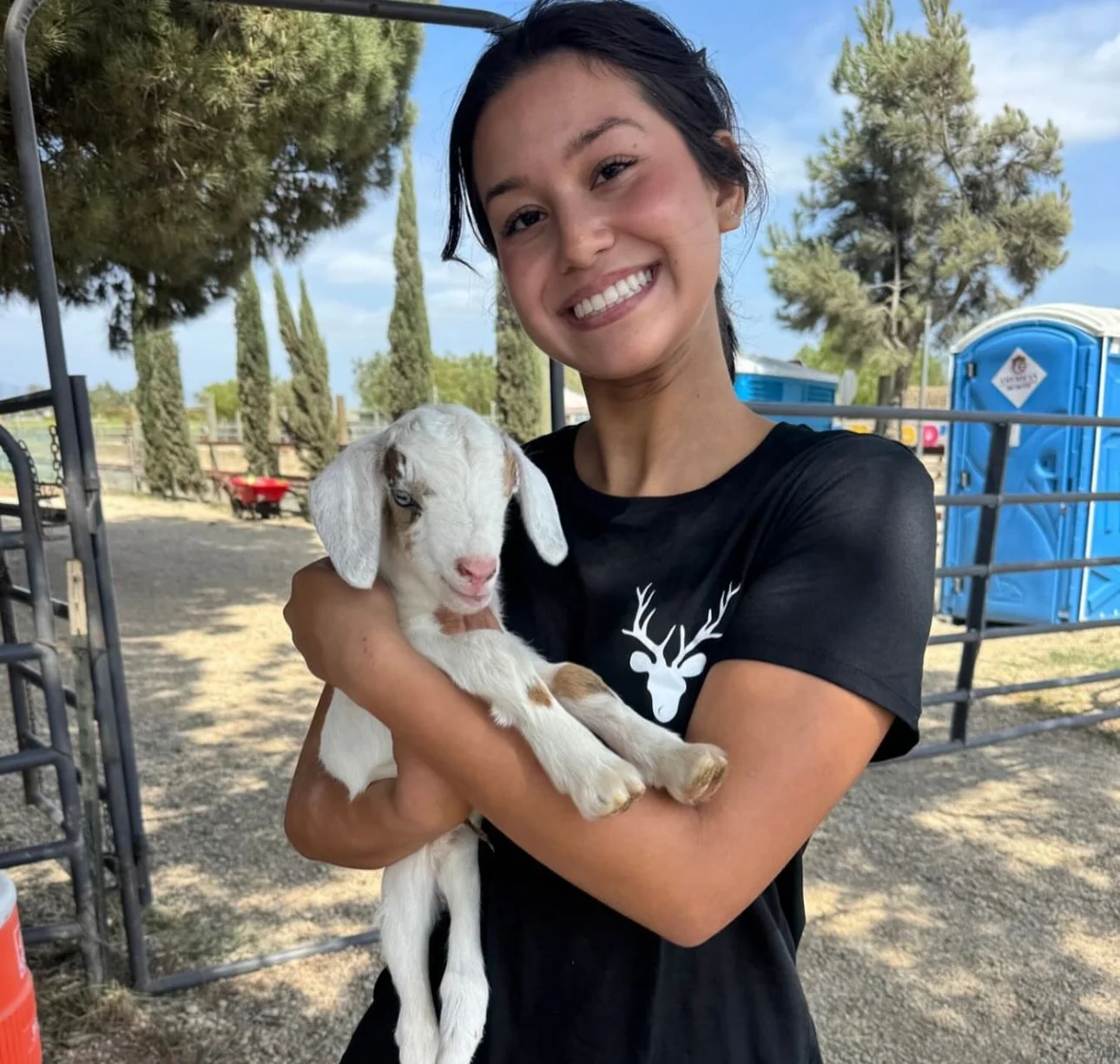 A woman smiling and holding a baby goat outdoors on a sunny day.