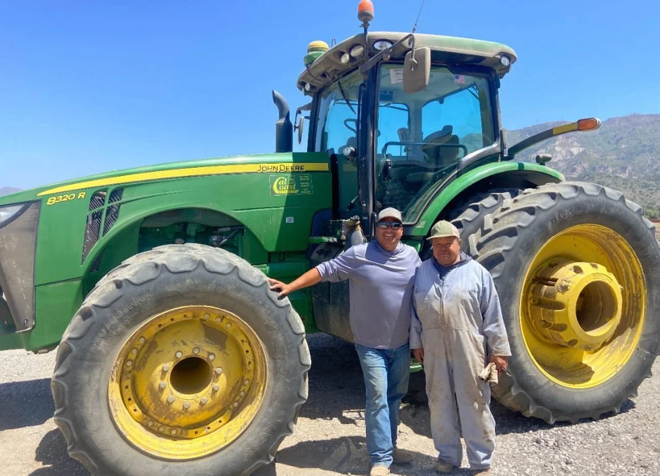 Two men standing in front of a large green John Deere tractor outdoors under a clear blue sky, with mountains in the background. One man is smiling and wearing sunglasses and a baseball cap, the other is wearing a work coat and a hat.