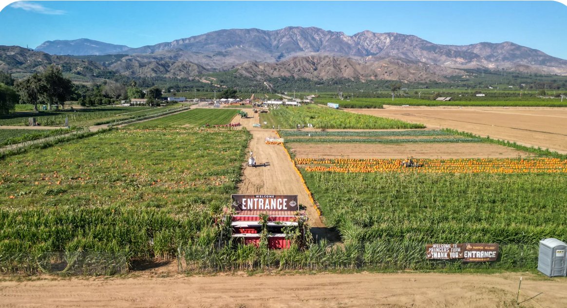 A scenic farm with various crops and a mountain range in the background, featuring a dirt path leading to the entrance with signs welcoming visitors.