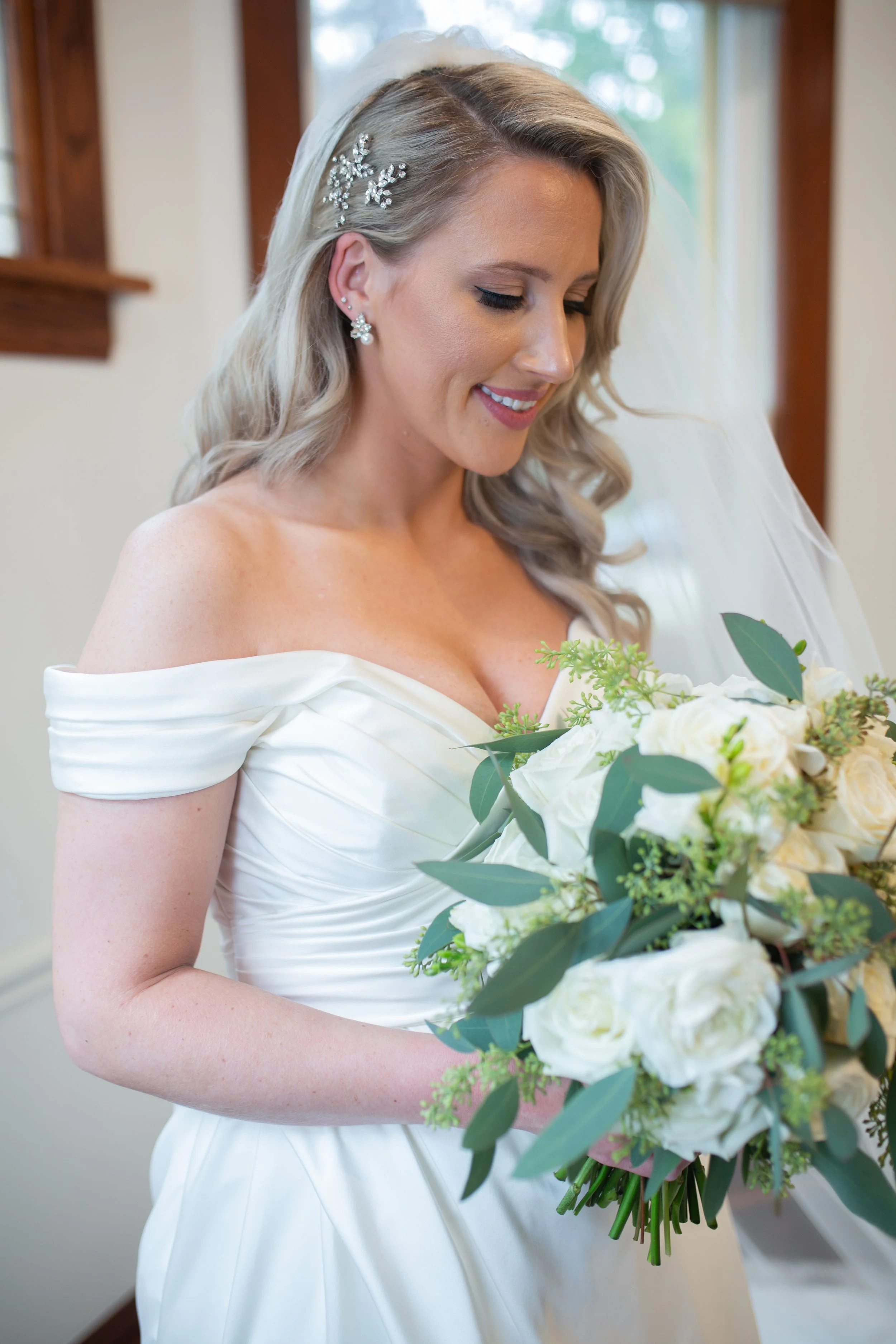 Close-up of a bride with beautiful makeup holding a bouquet of white roses and green foliage, wearing a white wedding dress, with blonde hair styled in loose waves and adorned with a silver hairpiece and earrings, smiling with eyes closed.