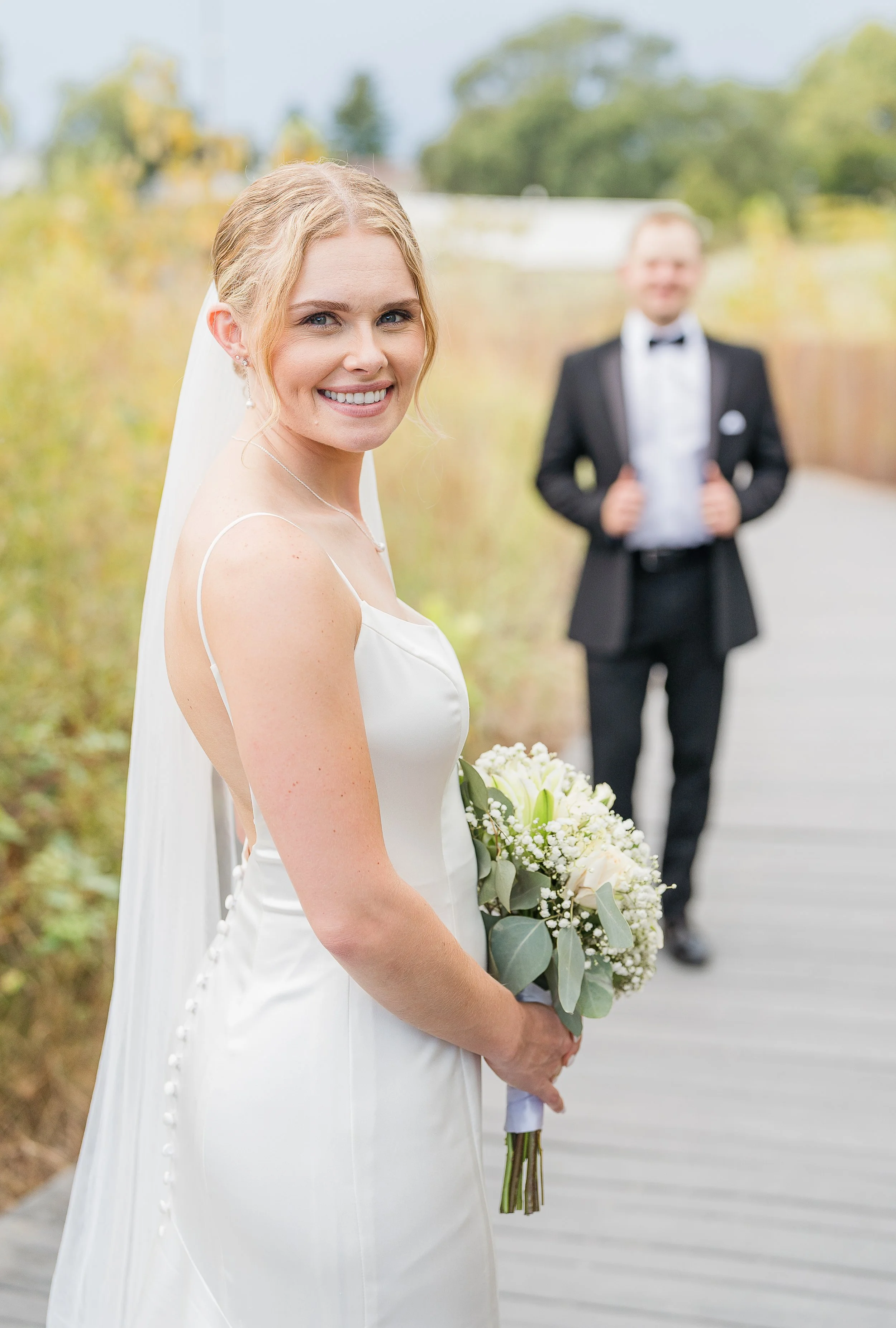 A bride in a white wedding dress and beautiful makeup holding a bouquet of white flowers and greenery, smiling, standing outdoors with a blurred groom in a tuxedo in the background.