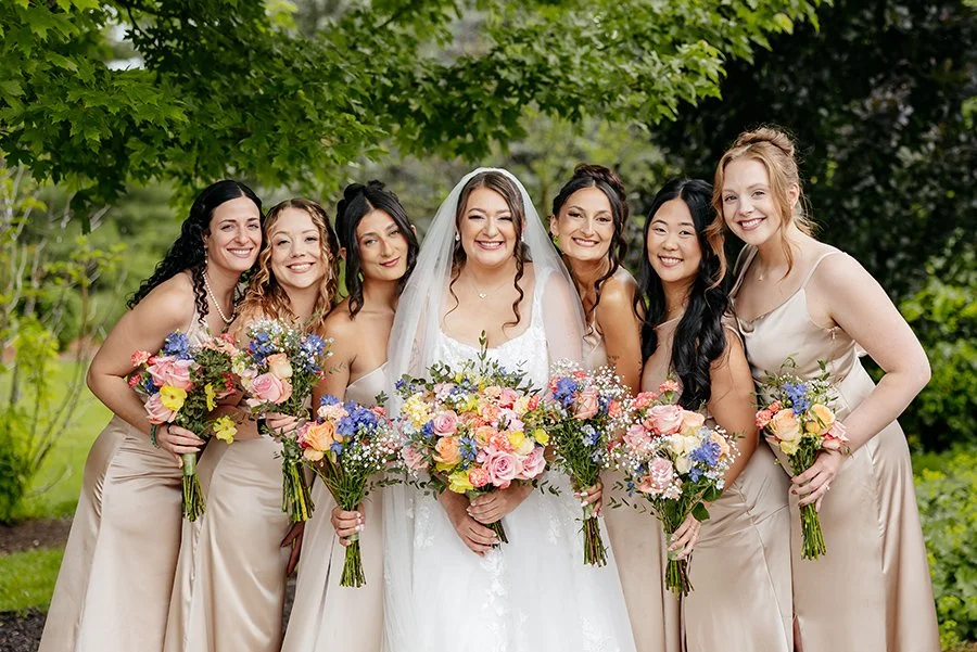 A bride with beautiful glam makeup in white wedding dress with her bridesmaids in beige dresses holding colorful bouquets, outdoors with green trees.