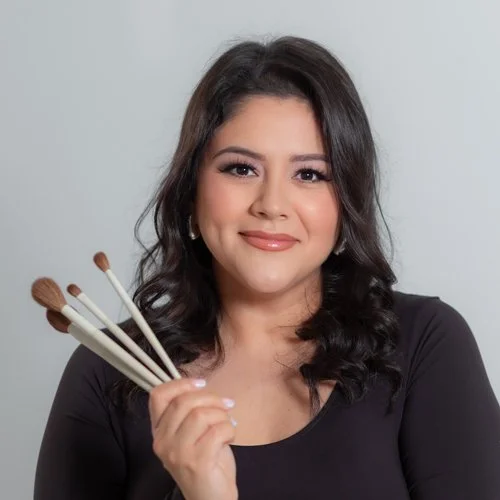 A female makeup artist with dark hair holding makeup brushes, smiling, wearing a black top, against a light background.
