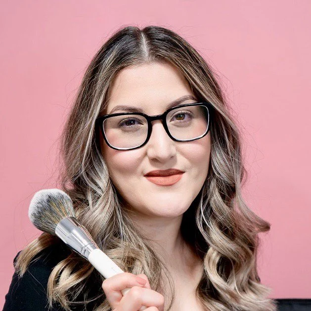 A makeup artist with wavy blonde hair wearing glasses and a black top, holding a makeup brush against a pink background.