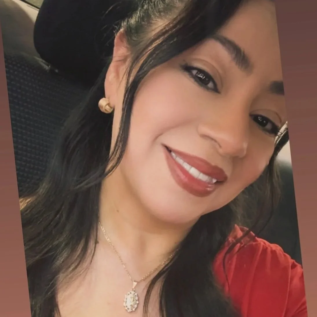 A dark-haired makeup artist with a glam makeup look wearing a red top, gold earrings, and a necklace with a pendant. 