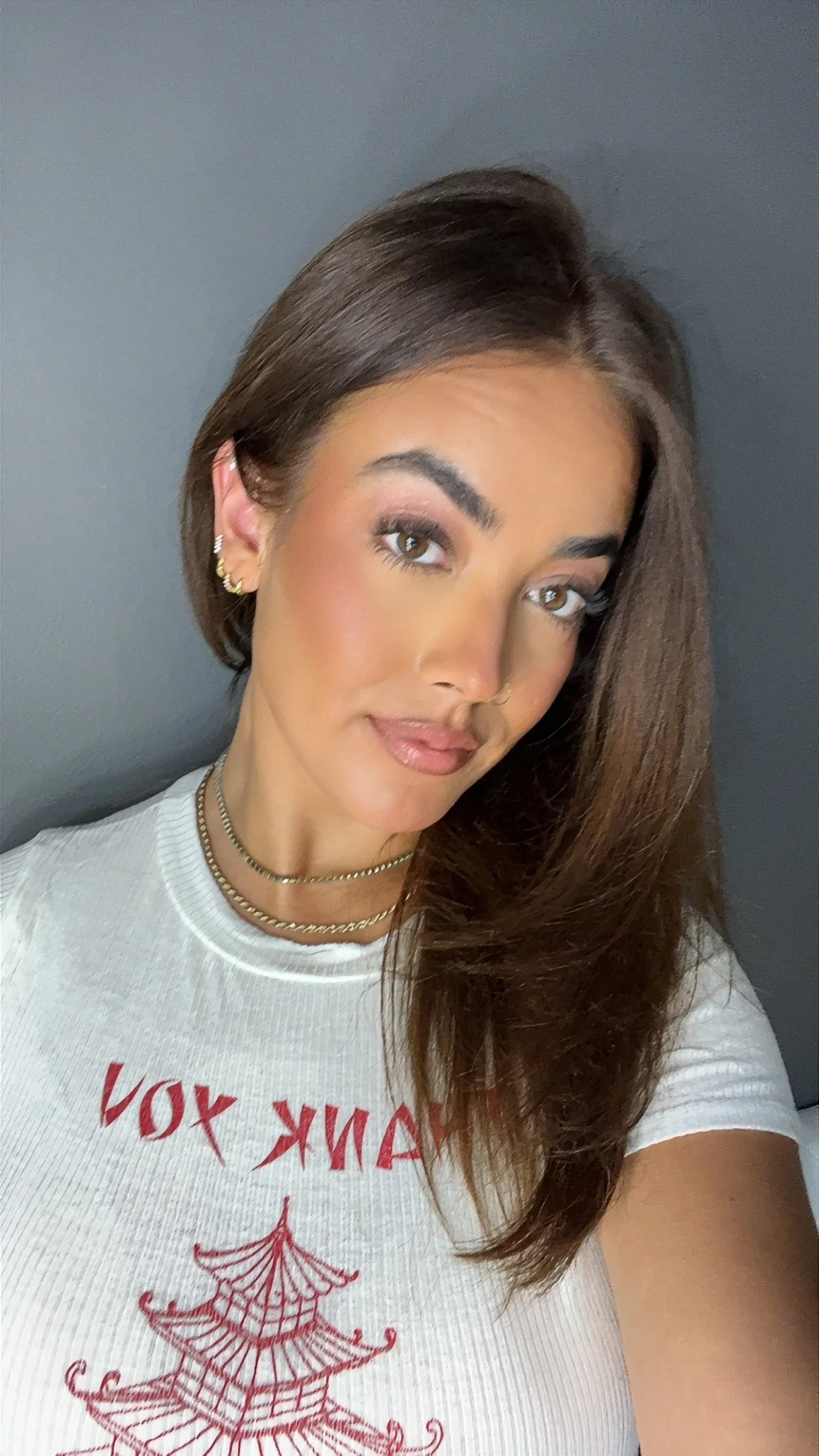 A hairstylist with shoulder-length brown hair, wearing earrings and layered necklaces, posing against a gray background. She is wearing a white t-shirt with red text and a graphic design.