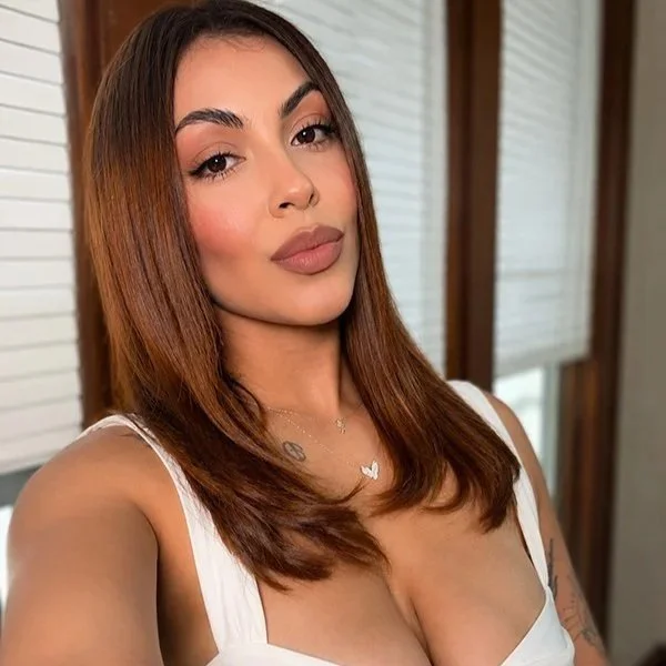 A hairstylist with glam makeup andstraight, shoulder-length brown hair taking a selfie indoors. She is wearing a white sleeveless top and has natural makeup with nude lipstick. Behind her are windows with blinds.