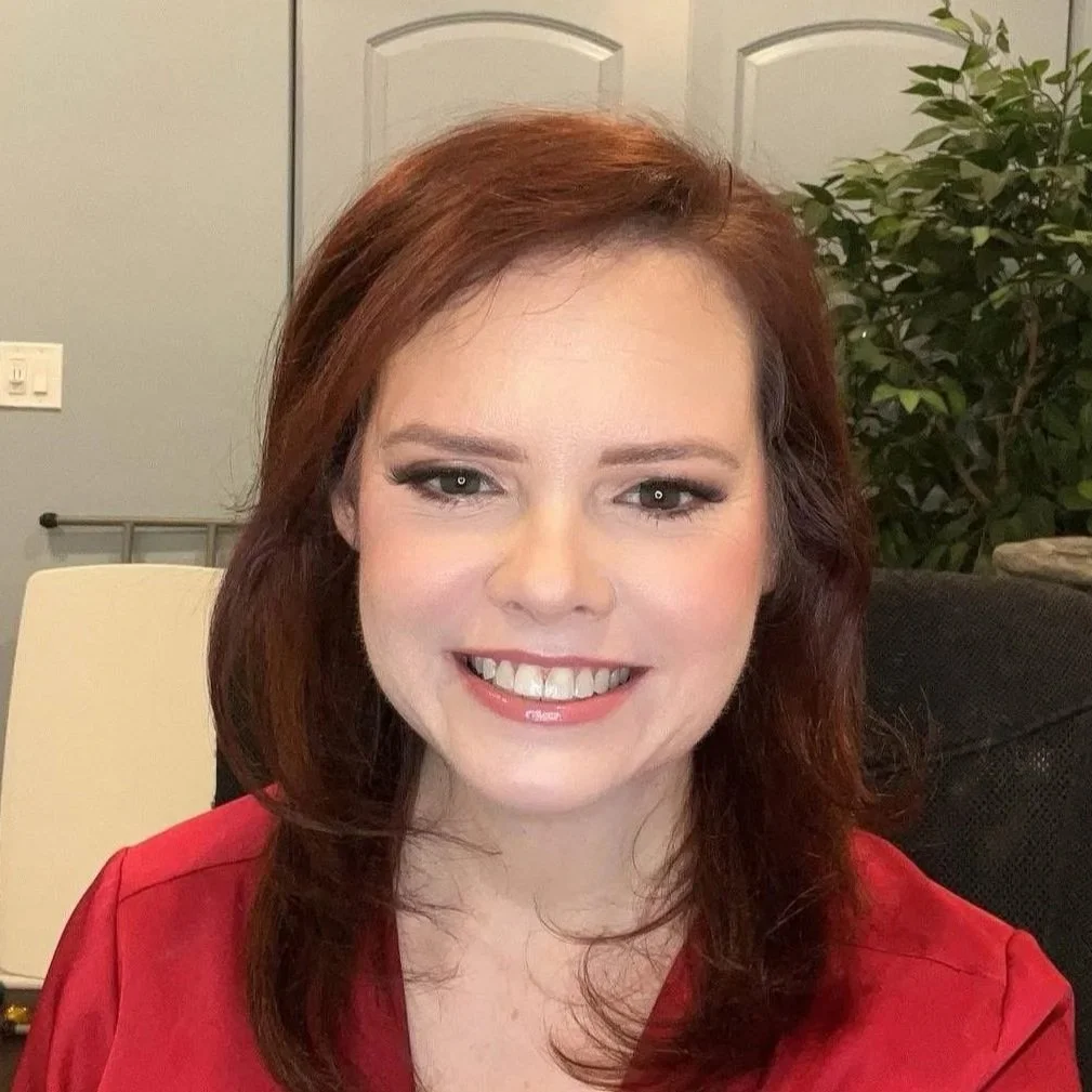 A young female makeup artist with shoulder-length auburn hair, wearing a red top, smiling, with a background of a door, a wall with light switches, and a plant.
