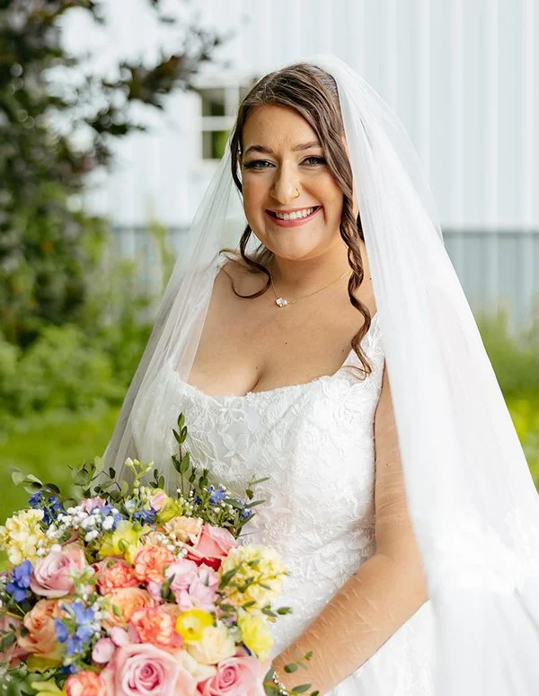 A smiling bride with soft glam makeup wears a white lace wedding dress and veil while holding a colorful bouquet of flowers outdoors.