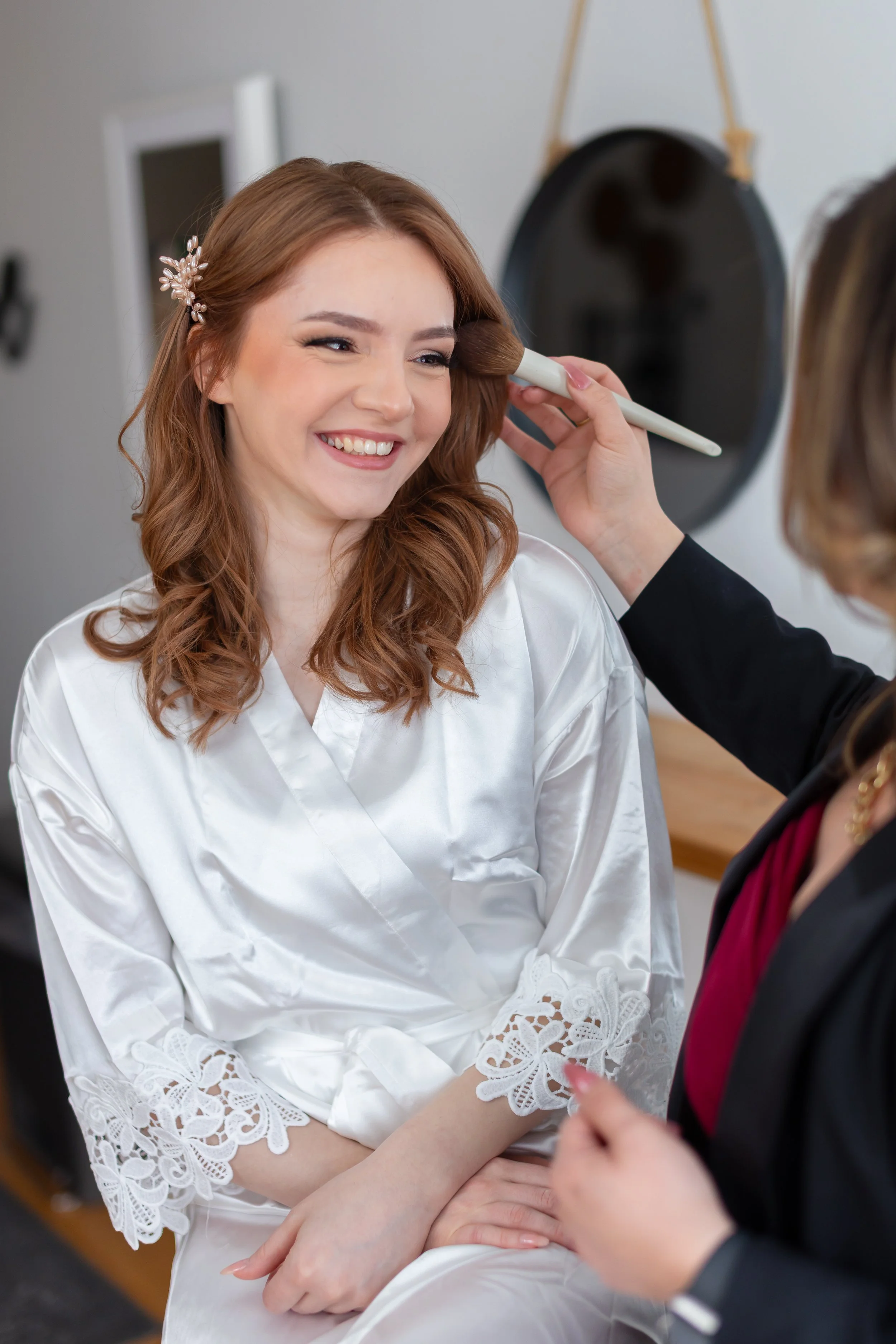 A bride with long, wavy auburn hair and a floral hairpin smiling as she gets her makeup done by professional make upartist Megan Orozco, who is partially visible, in a room with a round black wall hanging in the background.