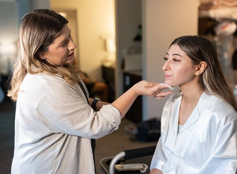 Makeup artist applying makeup to a woman sitting with her eyes closed in a cozy room.