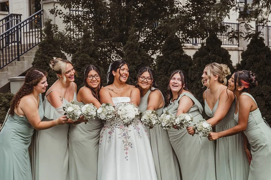 A bride and seven bridesmaids standing outdoors, each holding white bouquets and laughing after having their makeup professionally done by Glam Luxe Artistry.