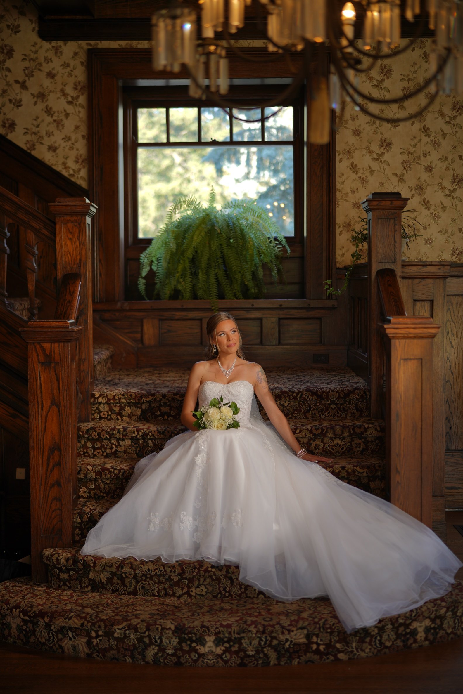 A bride in a white wedding dress with glam makeup and an updo hairstyle sitting on a staircase in a formal wood-paneled room, holding a bouquet of white roses, with a large window and greenery outside, and a chandelier hanging above.