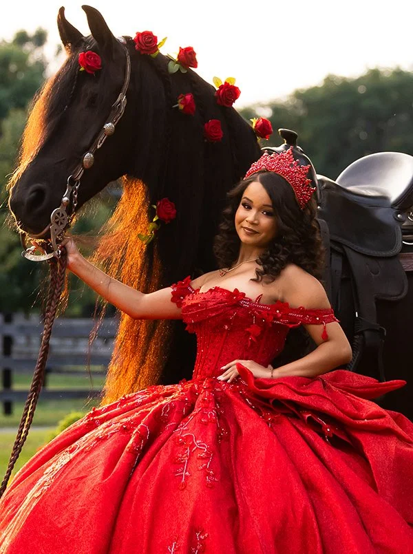 A young woman in a red Quinceañera dress and a matching tiara stands next to a black horse decorated with red roses in its mane. The girl is smiling at the camera, showing off her professionally applied glam makeup.