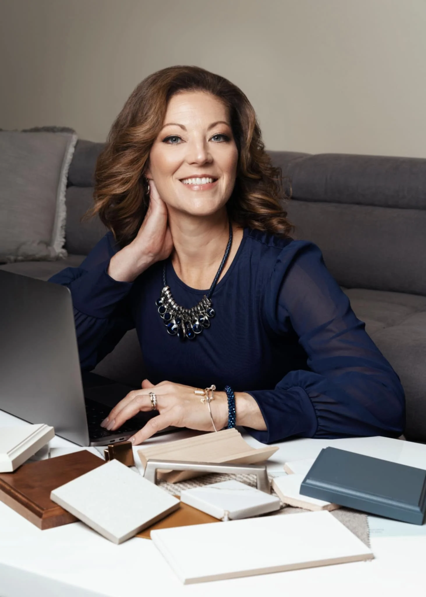 A professional business woman with brown wavy hair smiling at the camera, sitting at a desk with multiple sample books and fabric swatches, in a home or office setting having headshots taken by a photographer.