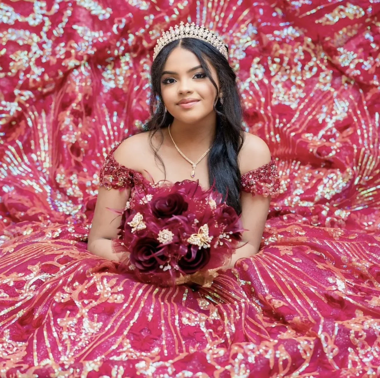 A young woman poses for a Quinceañera photo surrounded by the layers of her pink and gold dress, wearing a tiara and holding a bouquet.