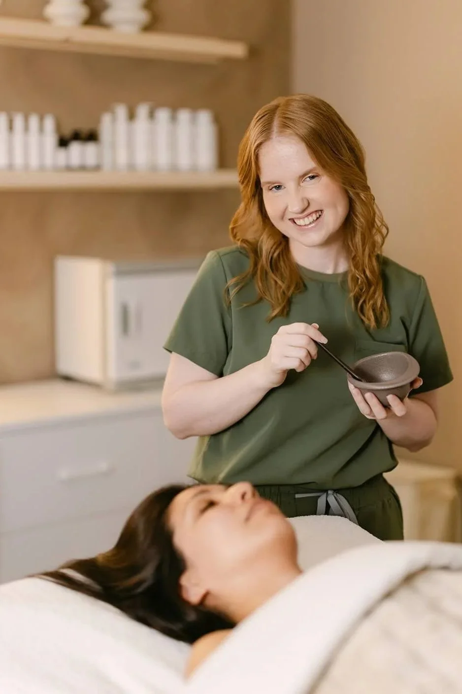 A female professional esthetician with professionally done makeup and red hair poses for branding photos while holding a bowl and a spoon near a patient lying on a bed.