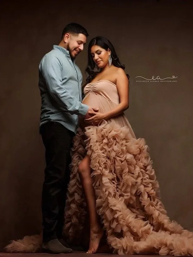 A pregnant woman with professionally applied glam makeup wears a ruffled, peach-colored gown standing next to a man in a blue shirt in a pregnancy photo pose.