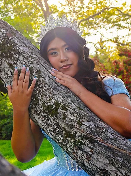 A young woman with beautiful glam makeup is celebrating a Quinceañera in a light blue dress wearing a crown, leaning on a tree trunk outdoors with greenery and sunlight in the background.