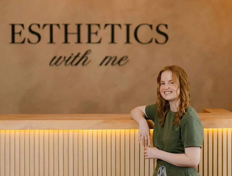 A woman with red hair in green scrubs smiles for branding photos while standing at a wooden reception desk in front of a wall that says 'ESTHETICS with me' in black letters.