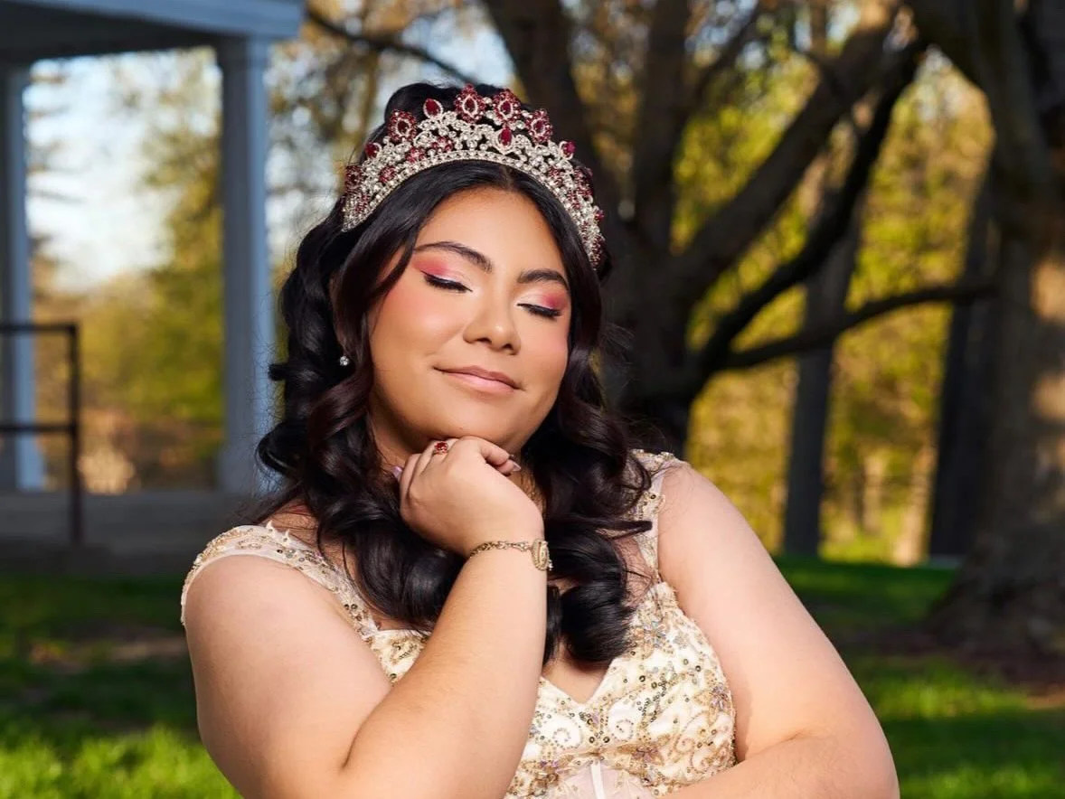 Young woman with a glam makeup look celebrating her Quinceañera wears a tiara and elegant dress, smiling with eyes closed outdoors in a park with trees and a pavilion in the background.