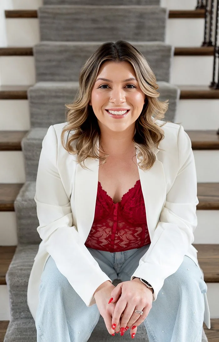 Professional makeup artist and owner of Glam Luxe Artistry, Megan Orozco smiles, wearing a white blazer, red lace top, and light blue jeans, sitting on stairs indoors.