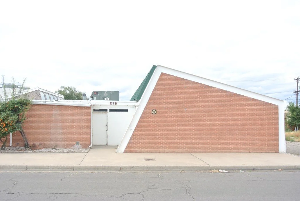 A modern building with a sloped brick wall and white framing, an entrance door, and sidewalk in front.