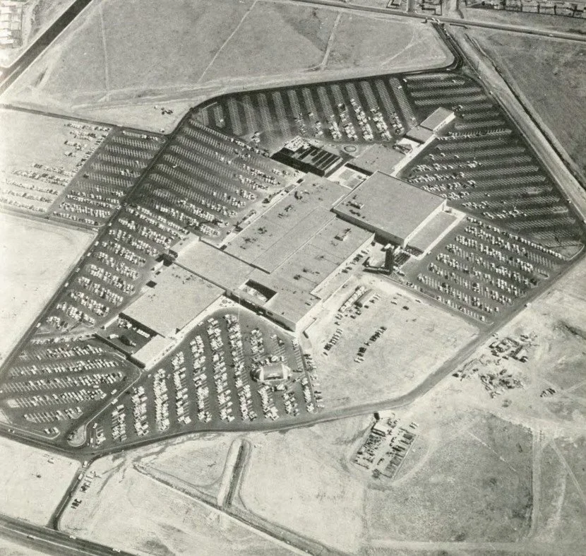Aerial view of a large sports arena surrounded by a parking lot, with sections of parked cars and open fields nearby.