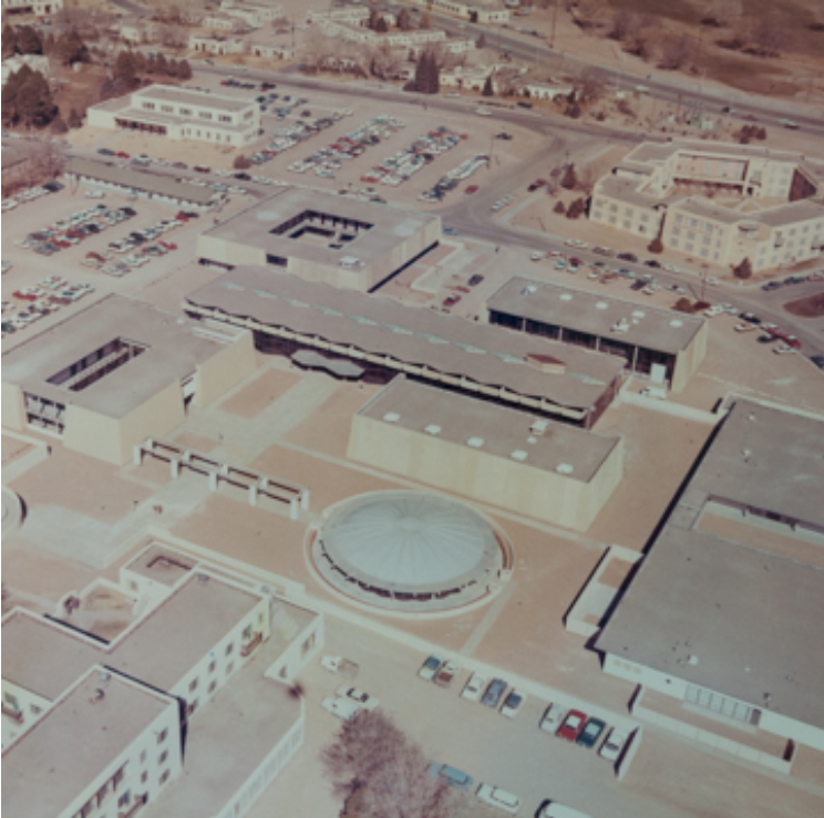 An aerial view of a school complex with multiple buildings, parking lots, and a large circular structure, surrounded by a desert landscape.