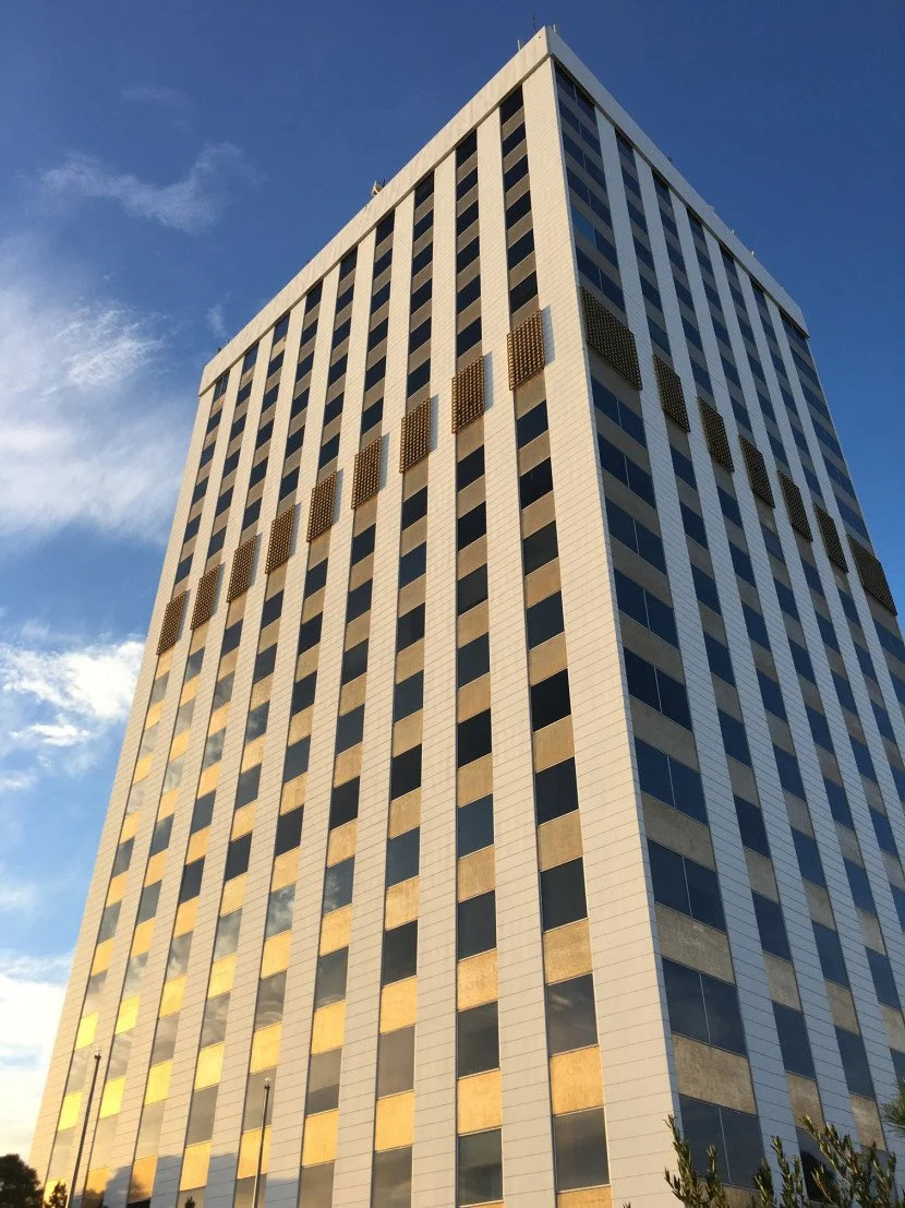 A tall modern office building with numerous windows reflecting the sky and clouds, under a clear blue sky.