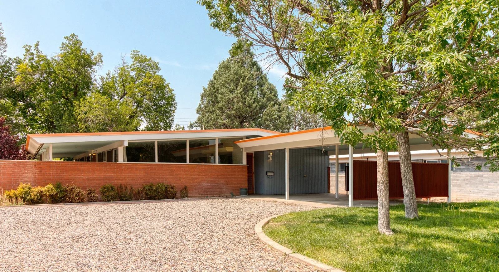 Mid-century modern house with a flat roof, large glass windows, a brick wall, and a gravel driveway, surrounded by green trees and lawn.