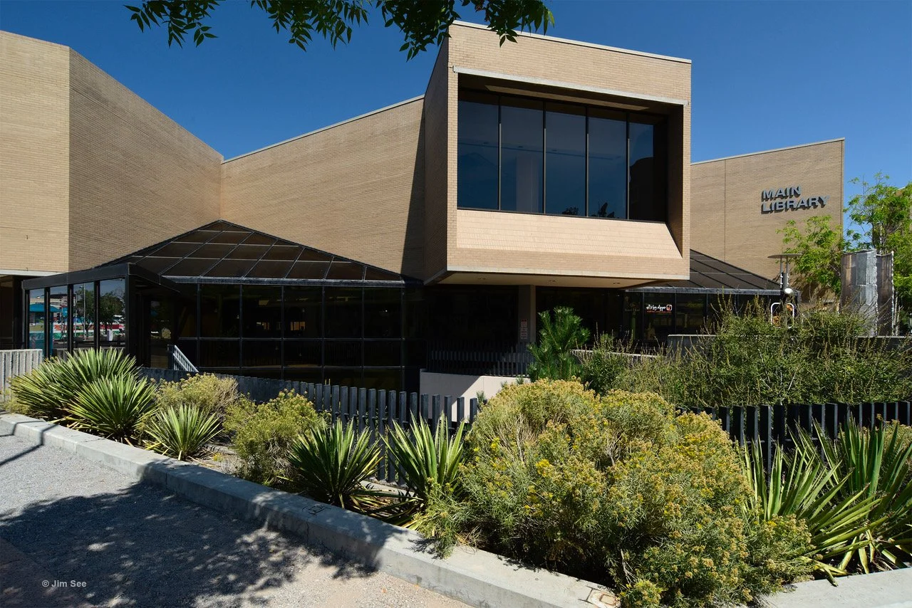 Main library building with modern architecture, beige brick walls, large glass windows, and surrounding desert plants under a clear blue sky.
