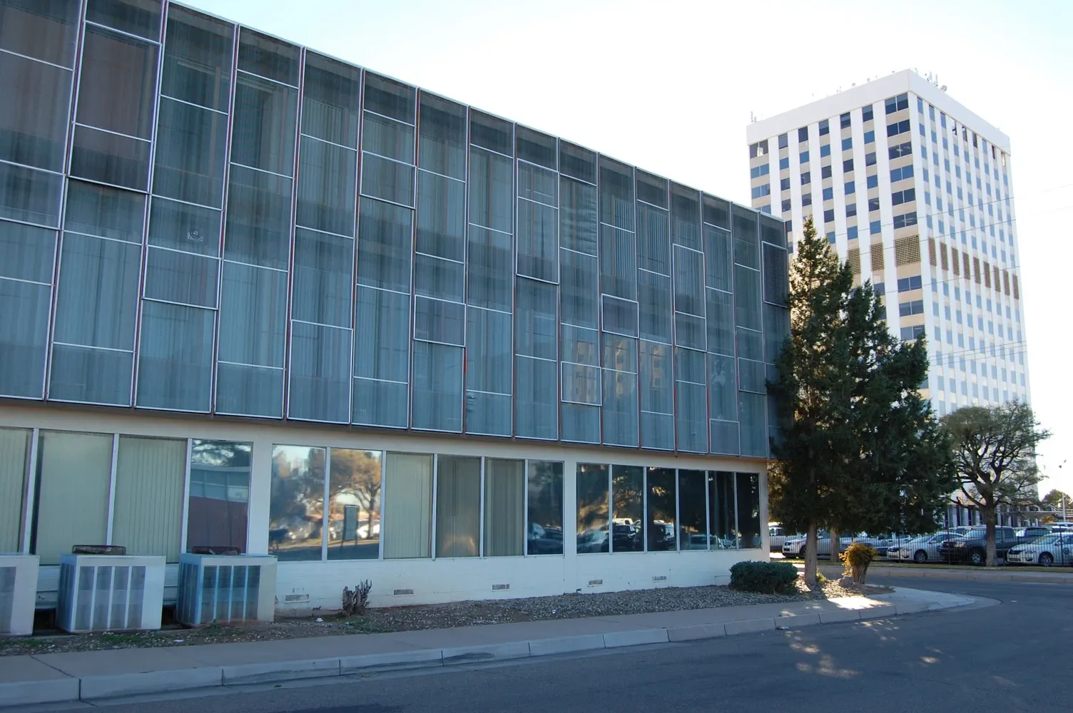 A city street scene with a modern, glass-fronted building in the foreground and a tall, white, grid-patterned office building in the background. There are trees and parked cars along the street.