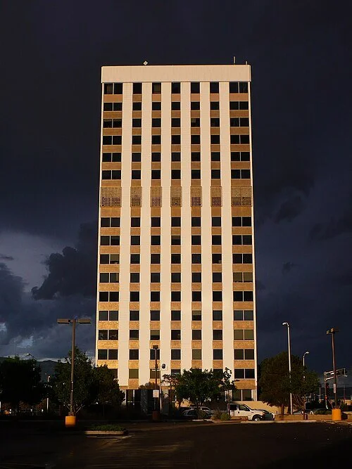 Tall office building with multiple floors and a grid of windows, situated in an urban area during dusk or evening.
