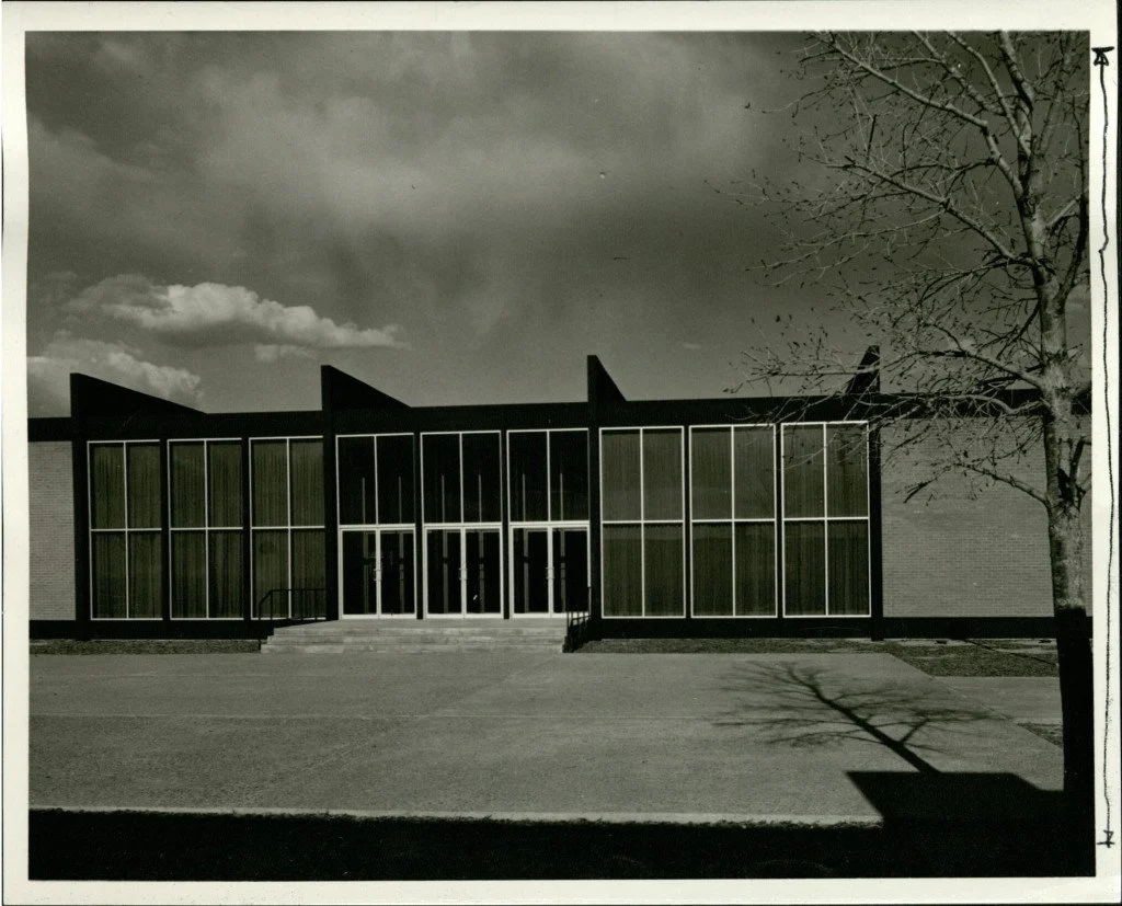 Black and white photo of a modern building with large glass windows and stepped entrance, with a leafless tree casting a shadow in the foreground.