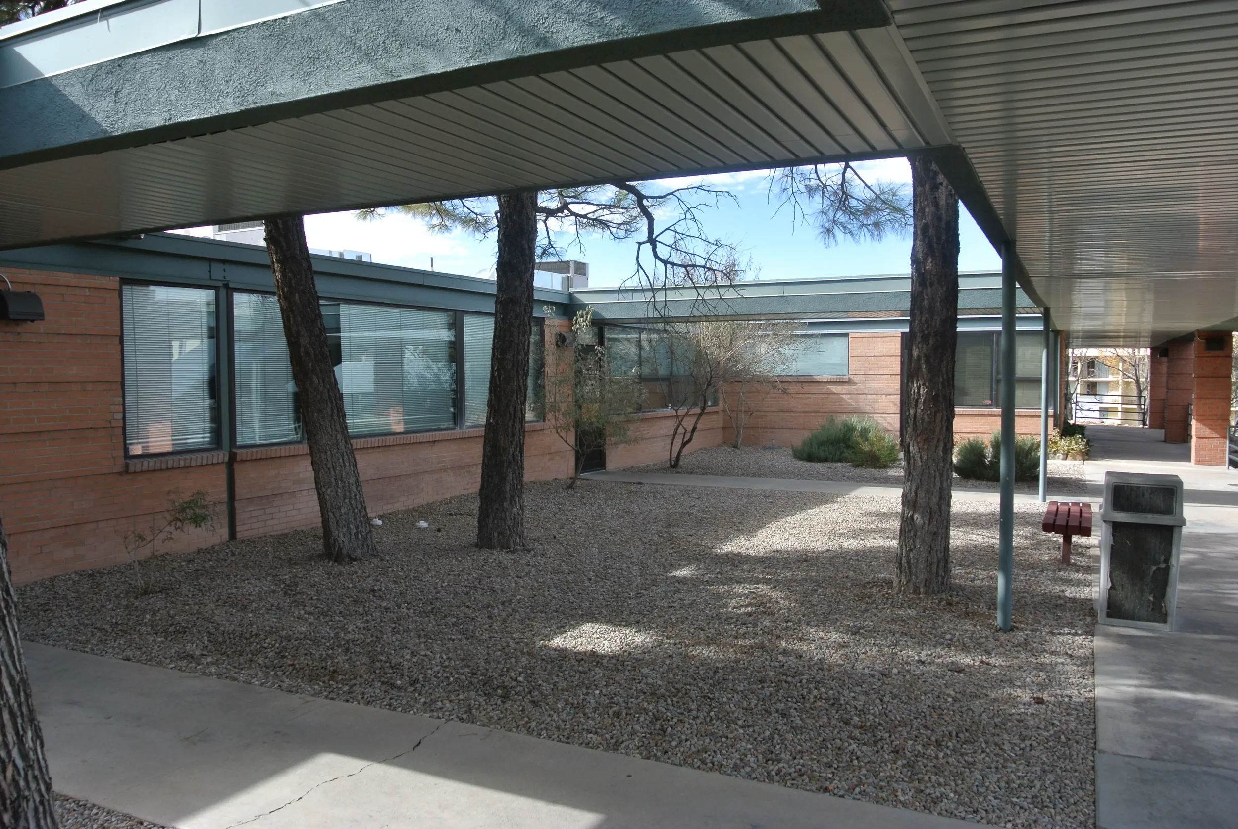 Exterior view of a modern building with large windows, surrounded by trees and a gravel landscape, under a metal roof structure.
