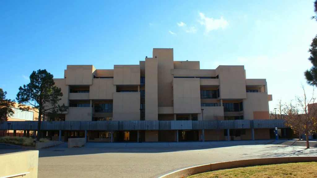 A multi-story beige condominium building with several setbacks, surrounded by trees and a paved parking area in the foreground, under a clear blue sky.